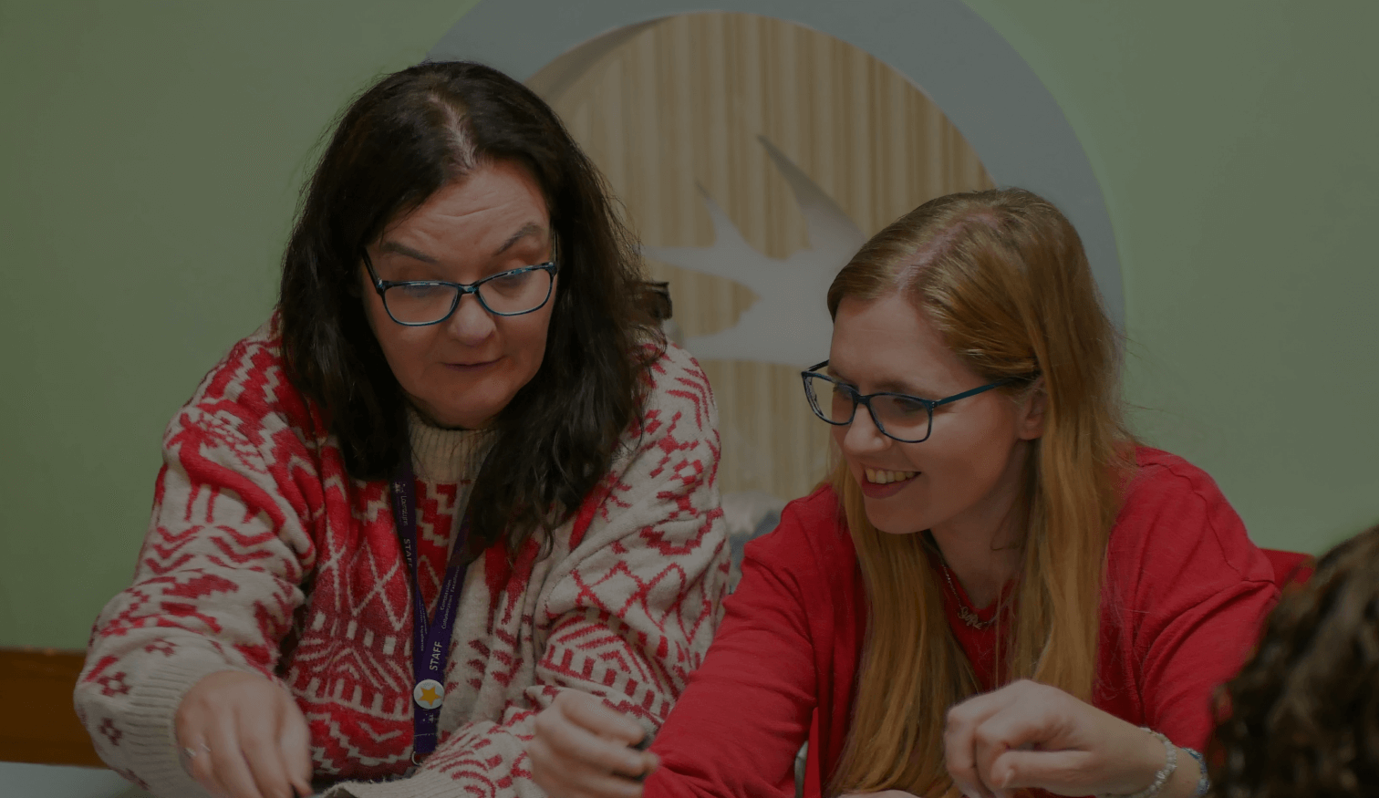 Two women wearing glasses sitting at a table, smiling and engaged in a creative or educational activity.