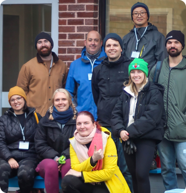 Group of adults dressed in warm outdoor clothing posing together and smiling outside a brick building.