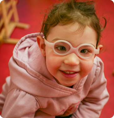 Smiling young girl in a light pink hoodie and white glasses, looking up at the camera against a red background.