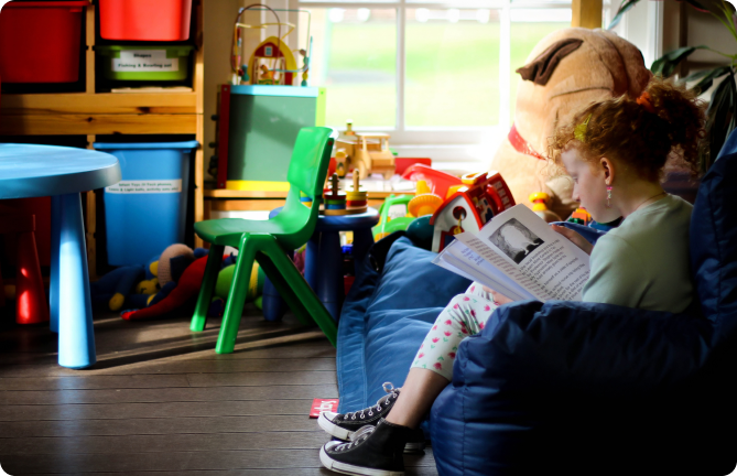 Young girl with curly red hair sitting on a blue beanbag, reading a book in a colourful playroom filled with toys and bright furniture.
