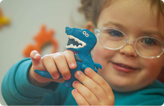 Young girl with glasses smiling and holding up a small blue dinosaur made from playdough.