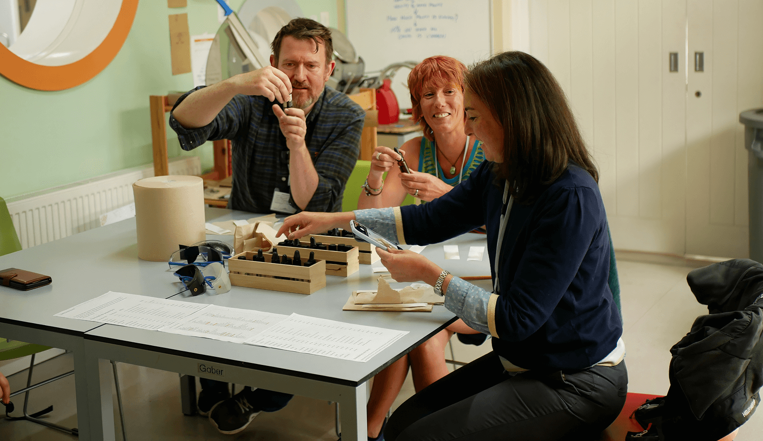 Three adults sitting at a table during a workshop, examining small dropper bottles. The two women are smiling while the man demonstrates one of the bottles.