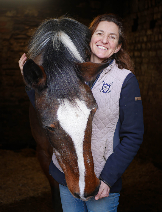 A smiling woman with curly brown hair, wearing a light quilted vest over a dark long-sleeve shirt, stands closely beside a brown horse with a distinctive white blaze on its face and a black mane with a white streak. They are in a dimly lit stable with stone walls. The woman gently holds the horse’s head.