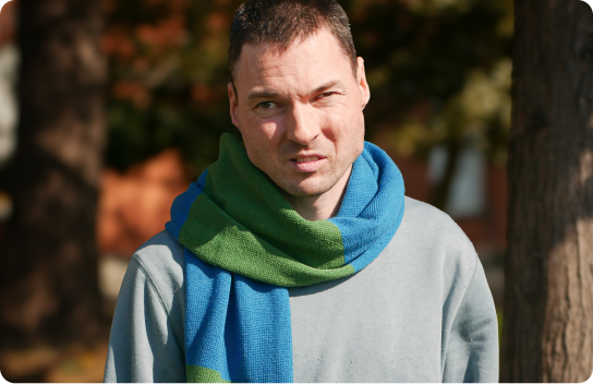 A man stands outside in sunlight with trees and a building in the background. He wears a gray sweater and a cozy scarf wrapped around his neck in blue and green colors.