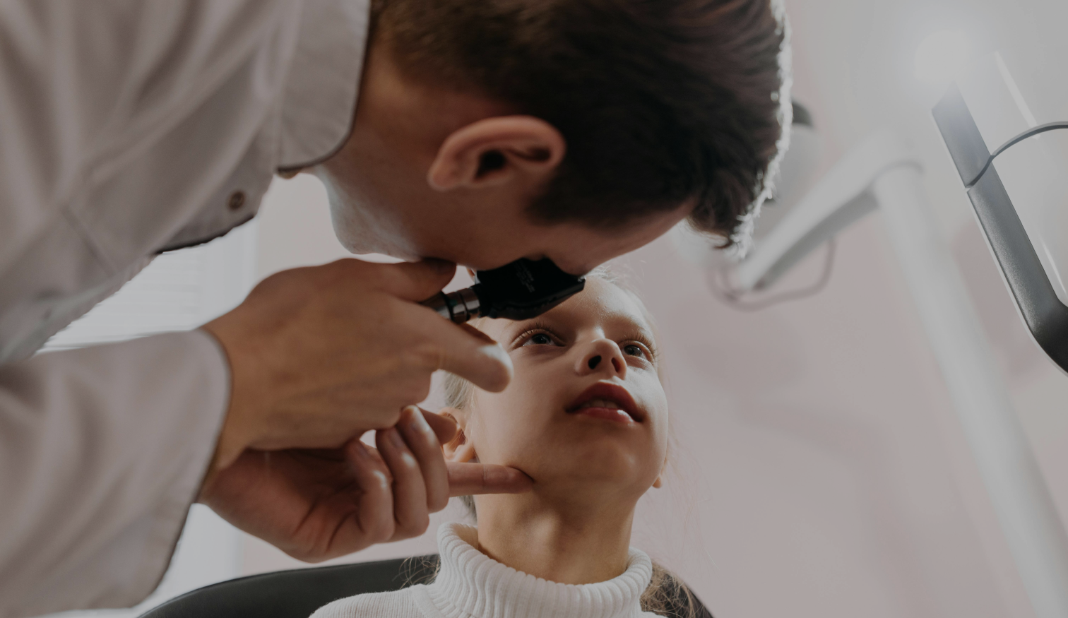 A young girl sits calmly while a male healthcare professional closely examines her eye using an ophthalmoscope. The girl is wearing a white turtleneck sweater and looking slightly upward as the examination takes place.