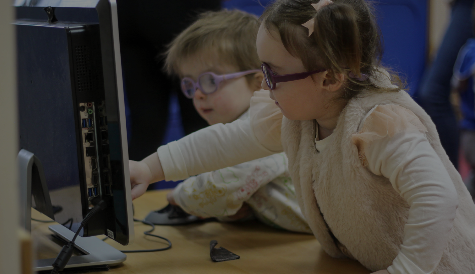 Two young girls wearing glasses using a desktop computer together, one pointing at the screen while the other holds the mouse.