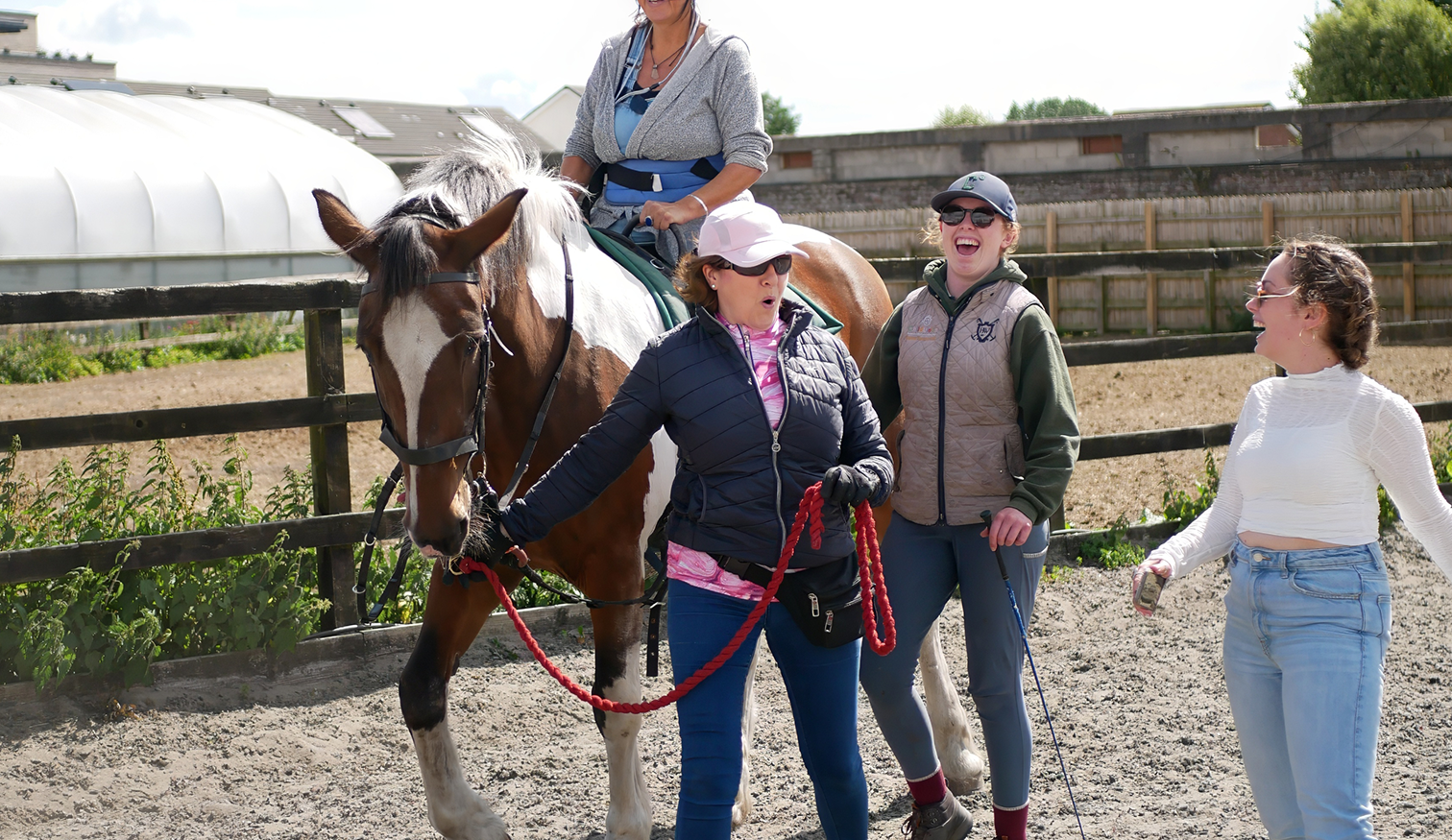 Group of women supporting a horse-riding session in an outdoor arena, with one woman riding and others walking alongside the horse, laughing and smiling together.