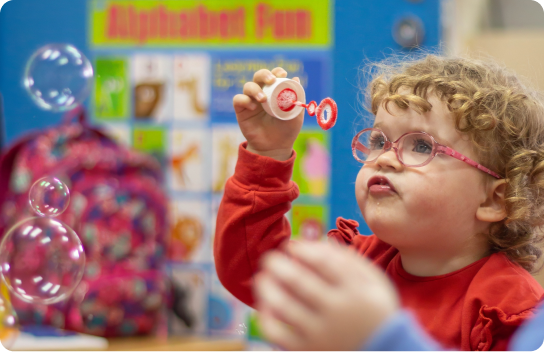 Young girl with curly hair and glasses blowing bubbles in a classroom, focused and engaged, with an “Alphabet Fun” chart visible in the background.