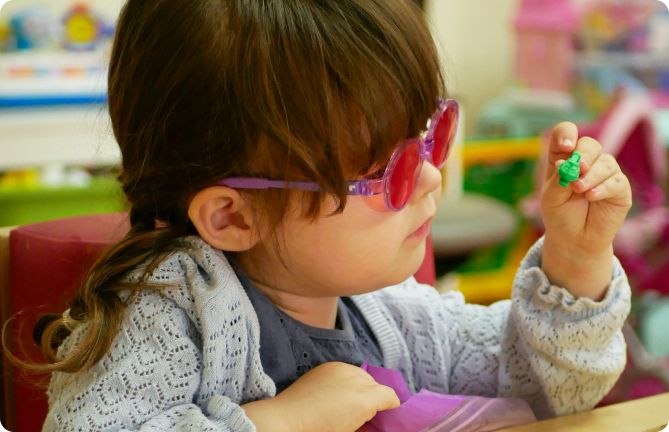 Young girl wearing pink-tinted glasses examining a small green object closely, seated at a table in a colourful classroom or play area.