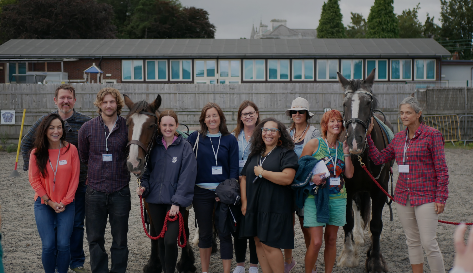 Group of adults smiling and posing with two horses in an outdoor paddock, all wearing name tags at an event.