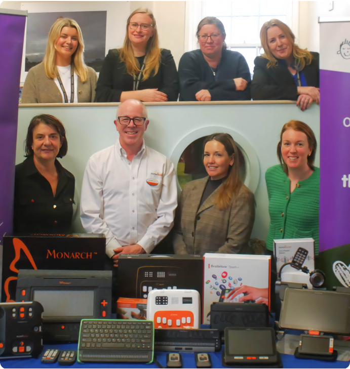 Group of adults standing behind a display table filled with assistive technology devices for people with visual impairments, including braille readers and tactile learning tools.