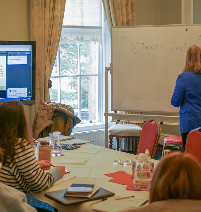Woman writing “Fossil fuels” on a whiteboard during a workshop or training session, with participants seated at a table taking notes. A screen displays related content beside a large window.