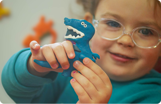 Young girl with glasses smiling and holding up a small blue dinosaur made from playdough.