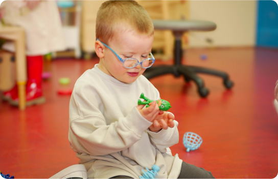 Young boy wearing a beige hoodie with blue rounded glasses playing with toys.