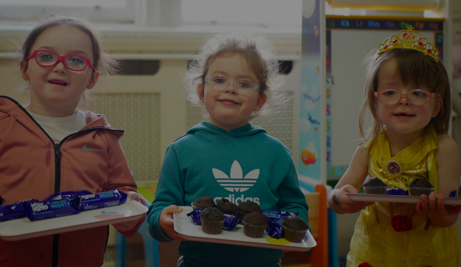 Three young girls smiling and holding trays with chocolate treats, standing in a classroom.