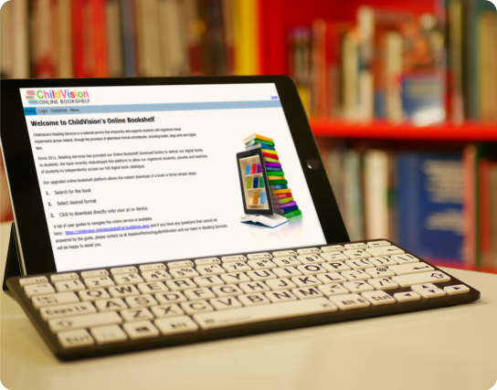 A tablet displaying ChildVision’s Online Bookshelf webpage is propped up on a table, with a large-key accessible keyboard in front. Bookshelves filled with colorful books are blurred in the background.