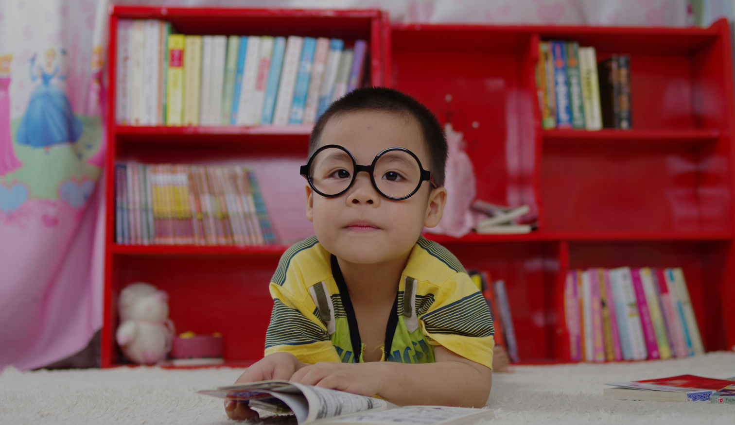 A young boy wearing round glasses lies on a soft carpet in front of a bright red bookshelf filled with colourful books. He looks directly at the camera while holding an open book in front of him.