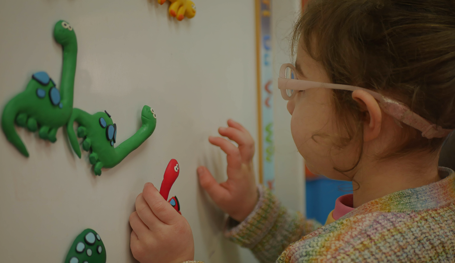A young girl wearing glasses and a hearing aid plays with colourful dinosaur shapes made from modeling clay, pressing them onto a whiteboard. She is focused and engaged in the activity.