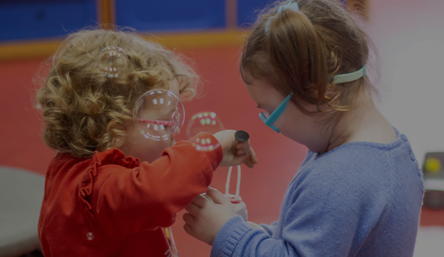 Two young girls wearing glasses, one in a red top and the other in a blue sweater, happily playing together with a bubble wand indoors.