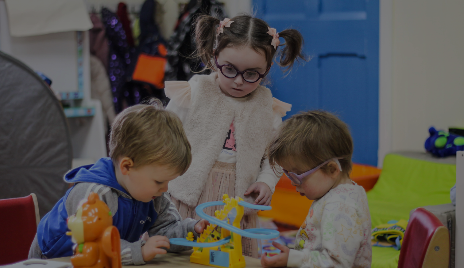 Three young children playing with toys indoors, two boys and one girl wearing glasses and a fluffy vest.